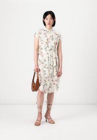 Blumenkleid aus weißem Stoff, kurze Ärmel, durchgehende Knopfleiste, tailliert mit Gürtel, mit bunten botanischen Mustern. Braune Handtasche und Riemchensandalen.