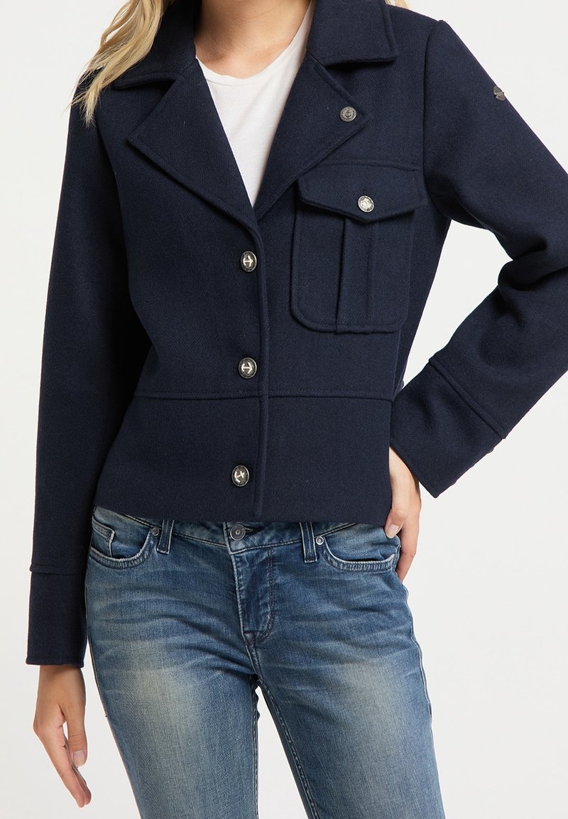 Žena v tmavě modré sako s knoflíky a kapsou na klopě, doplněná o světle modré džíny a bílé tričko.