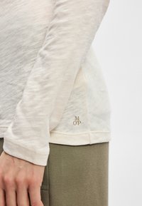Šviesiai smėlinė ilgesnė rankovėmis marškinėliai su tekstūruotu paviršiumi, su auksiniu "MOP" logotipu apatinėje dalyje ir suapvalinta apatine kraštine.