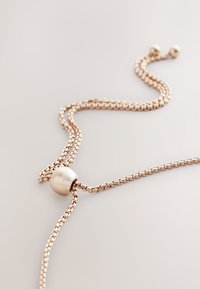 Two intertwined rose gold chains with a smooth round bead centerpiece. Chains are textured; the bead adds contrast in material.