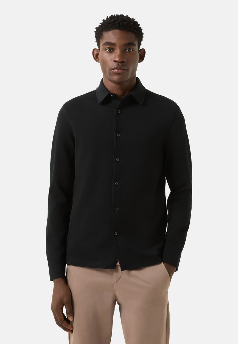 Schwarzes Hemd mit Knopfleiste aus weichem Stoff. Verfügt über lange Ärmel, einen klassischen Kragen und eine vordere Knopfleiste mit dunklen Knöpfen. Wird mit beigen Hosen getragen.