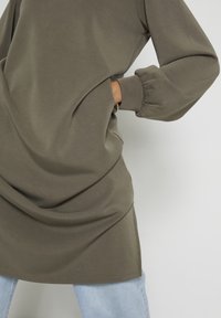 Zielono-oliwkowy, oversize'owy sweter z elastycznymi rękawami i kieszeniami po bokach; noszony na jasnoniebieskich dżinsach, wykonany z gładkiego, teksturowanego materiału.