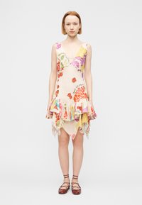 Νεαρή γυναίκα που φοράει αμάνικο φόρεμα με V-λαιμόκοψη και floral σχέδιο, με βολάν στο τελείωμα, και καφέ επίπεδα δερμάτινα παπούτσια με κορδόνια, στέκεται μπροστά σε απλό φόντο.