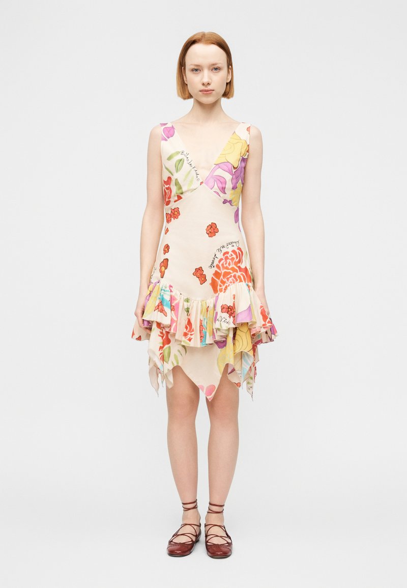 Νεαρή γυναίκα που φοράει αμάνικο φόρεμα με V-λαιμόκοψη και floral σχέδιο, με βολάν στο τελείωμα, και καφέ επίπεδα δερμάτινα παπούτσια με κορδόνια, στέκεται μπροστά σε απλό φόντο.