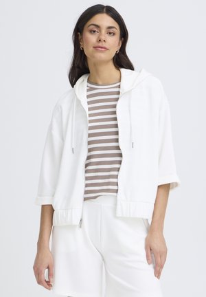 Kvinde med langt mørkt hår iført hvid hættetrøje med lynlås over beige og hvidstribet skjorte og hvide bukser, stående mod en ensfarvet baggrund.