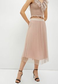Mujer con una falda midi plisada en rosa empolvado, una blusa sin mangas con volantes a juego y sandalias de tacón alto negro con tira al tobillo.