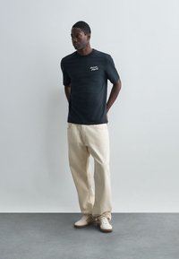 T-shirt preta de algodão com logo branco, combinada com calças largas bejes claras. Modelo a usar ténis casuais cremes, de pé contra um fundo neutro.