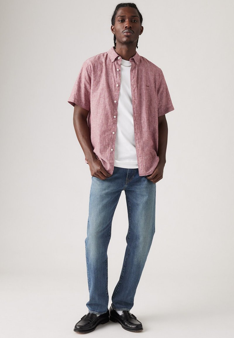 Chemise rouge à manches courtes avec un motif texturé, portée sur un t-shirt blanc, associée à un jean bleu et des chaussures noires.