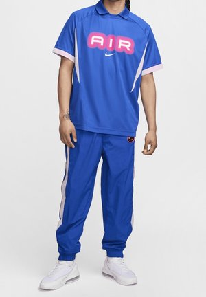 Persona con ropa deportiva azul con detalles en rosa y blanco, zapatillas blancas y un tatuaje en el antebrazo izquierdo.