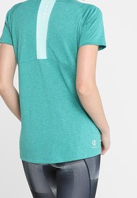 Tyrkysové sportovní tričko s krátkým rukávem, texturovaným zadním panelem a malým logem na spodním lemu, nošené s vzorovanými legínami.