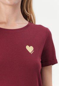 Žena nosí bordové tričko s malým zlatým srdiečkom a nápisom "ONLY" na ľavej strane hrudníka, krátke rukávy s obrubami.