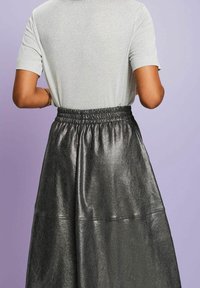 Femme portant une jupe taille haute gris foncé brillante avec une ceinture élastique et un haut léger à manches courtes, debout devant un fond violet clair.