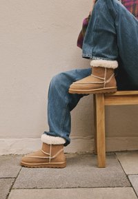 Botte en daim beige avec doublure en fourrure blanche, détail chaîne en métal sur le devant et semelle en caoutchouc texturée, portée avec un jean en denim retroussé.