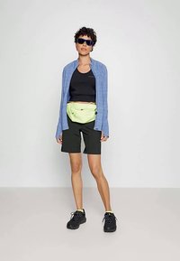 Ungdom som har på seg svarte treningsshorts og crop top, blå glidelåsjakke, neongul midjeveske, svarte joggesko og solbriller, står mot en ensfarget bakgrunn.