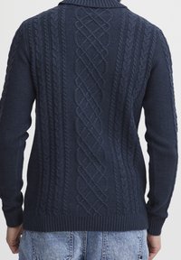 Pull en tricot torsadé bleu marine porté par une personne en jean en denim bleu clair, vu de dos.