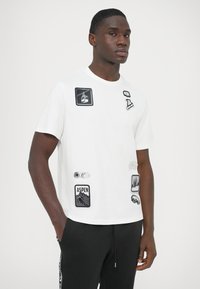 Biała krótka koszulka z krótkim rękawem z różnymi czarnymi i kolorowymi naszywkami, w tym z grafikami górskimi i narciarskimi, o okrągłym dekolcie.