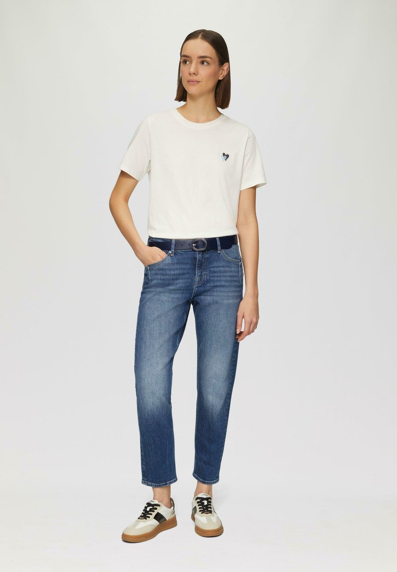 Witte T-shirt met korte mouwen met een hartgrafiek, blauwe spijkerbroek en sneakers met crèmekleurige, zwarte en tan accenten. Casual outfit.
