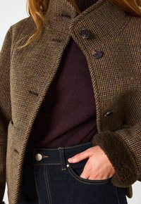 Giacca in misto lana a motivo houndstooth marrone con colletto alto, bottoni doppi e polsini in morbido pile marrone. Indossata con un maglione scuro e jeans.