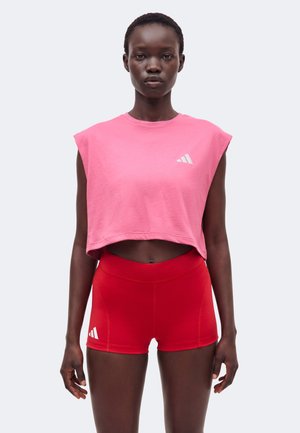 Femeie purtând un top roz fără mâneci și pantaloni scurți sport roșii, potriviți, stând în fața unui fundal simplu, deschis la culoare.