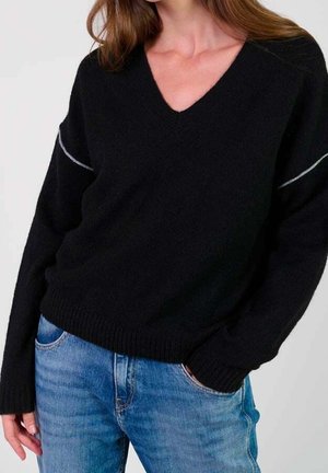 Femme portant un pull noir à col en V avec un détail subtil de couture blanche sur les manches et un jean bleu clair, debout avec une main dans la poche.