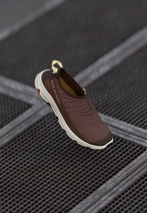 Pantof brown fără șireturi, cu un material texturat pe partea superioară și o talpă mai deschisă ca nuanță, având un element de prindere și un branding discret pe lateral.