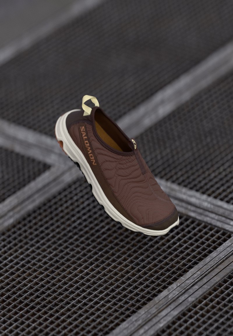 Rjavi natikač z teksturirano površino in kontrastnim svetlejšim podplatom, opremljen z zanko za poteg ter subtilnim logotipom ob strani.