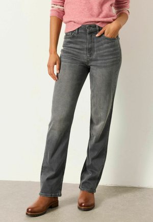 Vrouw draagt vervaagde zwarte jeans met rechte pijpen, bruine leren klompen en een roze trui met opgerolde mouwen.