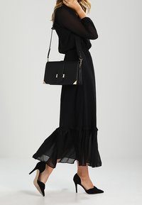 Robe maxi en chiffon noir avec un ourlet froncé, associée à un sac à main structuré noir avec des accents dorés et des talons noirs.