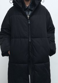 Černý péřový kabát s kapucí, zipem a bočními kapsami. Obsahuje prošívaný design a hladký povrch.