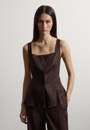 Frau mit langen dunklen Haaren, die ein ärmelloses, dunkelbraunes, mit Knöpfen versehenes Oberteil und passende Hosen trägt, vor einem einfarbigen hellen Hintergrund.