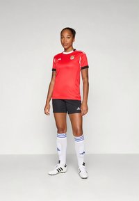 Czerwona koszulka piłkarska z czarnymi akcentami i logo, czarne spodenki, białe skarpety z niebieskimi paskami oraz czarne korki. Gładki, sportowy krój dopasowany do sylwetki.