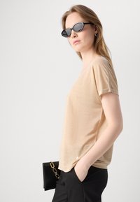 Beige top met korte mouwen in een zachte, gladde textuur, met een ontspannen pasvorm en opgerolde manchetten, gecombineerd met zwarte ovale zonnebril en zwarte clutch.