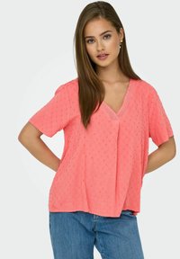 Koraalkleurig T-shirt met korte mouwen, een V-hals en een textuurpatroon van stippen. De stof is lichtgewicht met een relaxte pasvorm, gecombineerd met een blauwe spijkerbroek.