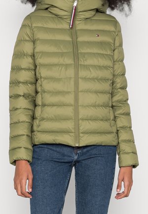 Persoon draagt een olijfgroene gewatteerde jas met ritssluiting en capuchon, gecombineerd met een blauwe spijkerbroek, staand tegen een effen achtergrond.