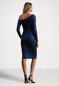 Φόρεμα σε ναυτικό μπλε χρώμα, έξω από τον ώμο, με μακριά μανίκια, στενή εφαρμογή, κατασκευασμένο από γυαλιστερό υλικό. Διαθέτει μήκος μέχρι το γόνατο και μαύρα πέδιλα με ψηλό τακούνι.