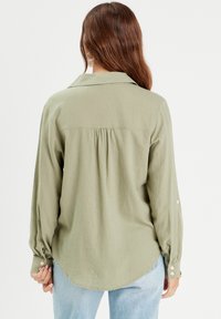 Chemise olive taille longue à manches longues en tissu doux, dotée d'un empiècement au dos et de détails froncés, avec des accents de boutons sur les poignets.