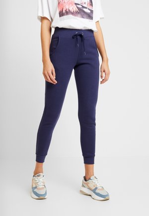 Vrouw in marineblauwe joggers met een drawstring taille en zakken, een wit grafisch T-shirt en blauwe, roze en witte sneakers.