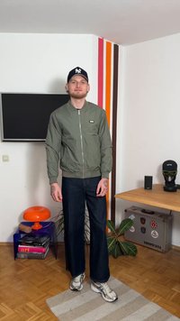 Maslinasto zelena bomber jakna s rebrastim manšetama, tamnoplave široke traperice i srebrne tenisice, smješteni u svijetlo uređenom unutarnjem prostoru.