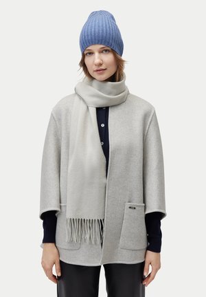 Frau mit einer blauen Strickmütze, einem hellgrauen Schal mit Fransen, einem hellgrauen Mantel mit Taschen, schwarzen Hosen und einem dunklen Hemd darunter.