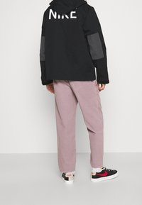 Black jacket with a hood and large white "NIKE" logo, gray accents on sleeves, paired with light pink pants and black sneakers with red detail.