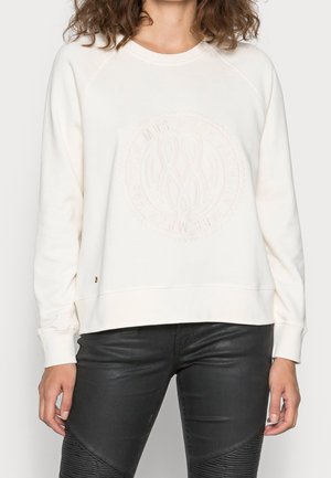 Kvinde iført en off-white langærmet sweatshirt med præget cirkulært logo og sorte teksturerede bukser.