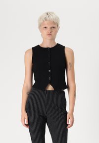 Μαύρο κοντό αμάνικο τοπ με στρογγυλή λαιμόκοψη, έξι κουμπιά και ελαφρώς καμπυλωτό στρίφωμα, σε συνδυασμό με παντελόνι με άσπρες και μαύρες ρίγες.