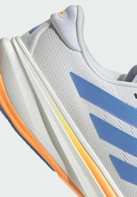 Primer plano de una zapatilla deportiva blanca con rayas azules y suela acentuada en naranja, mostrando la tela texturizada y el cuello del talón acolchado.