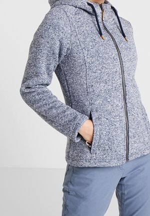 Persona che indossa una felpa con cappuccio e zip blu-grigio con tasche laterali e pantaloni azzurri, mano in tasca, su sfondo neutro.