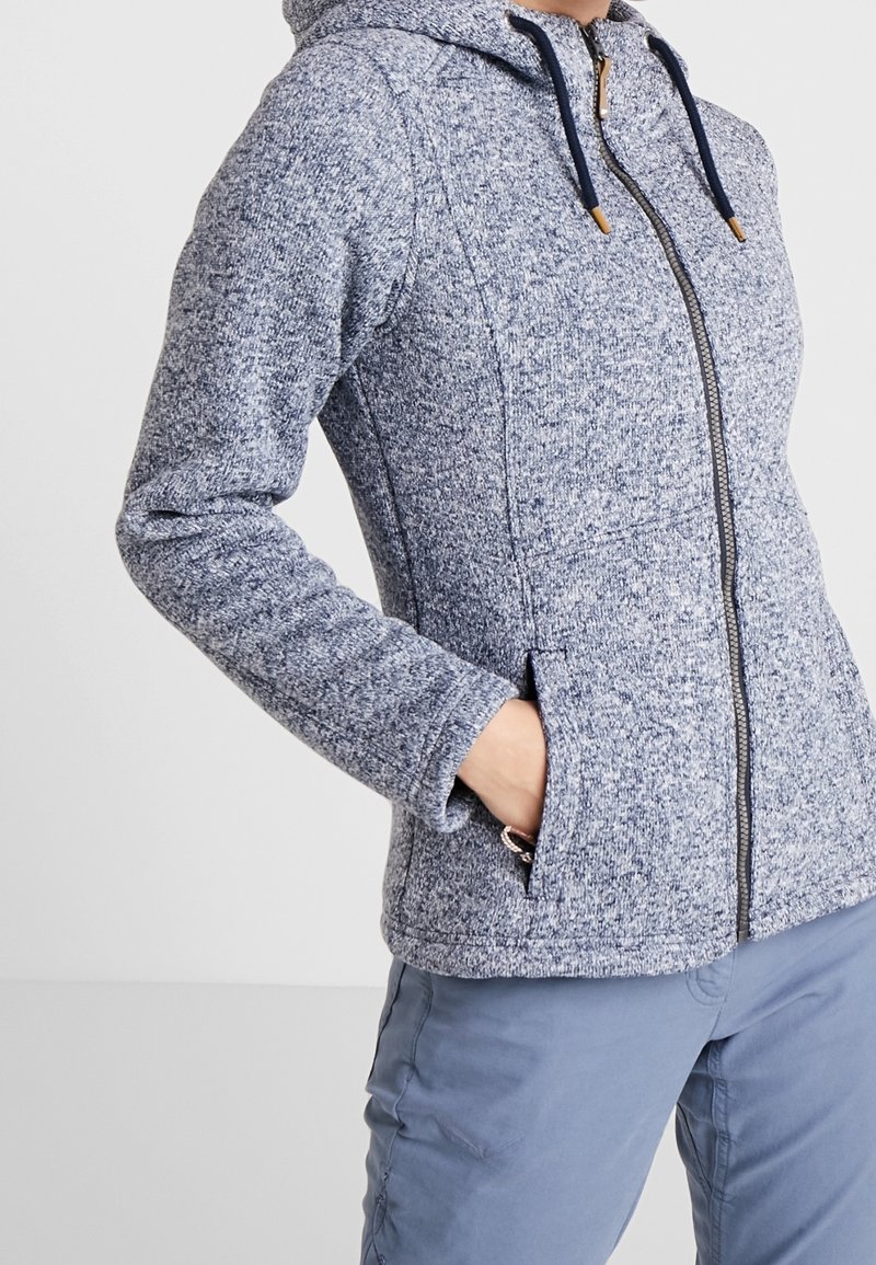 Personne portant un sweat à capuche zippé bleu-gris avec poches latérales et pantalon bleu clair, main dans la poche, sur fond uni.