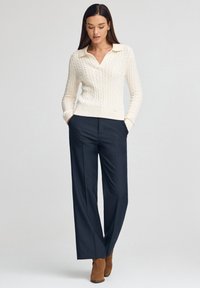 Cremefarbener Pullover mit Zopfmuster, V-Ausschnitt und umgeschlagenem Kragen, kombiniert mit maßgeschneiderten marineblauen Hosen und braunen Wildleder-Stiefeletten.