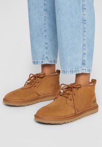 Bottes cheville en daim beige avec un bout arrondi, un design à lacets et une semelle en caoutchouc. Dotées d'une doublure intérieure douce et du logo UGG sur le côté.