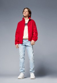 Rød lett jakke, beige sweatshirts med logo, lyseblå jeans og hvite sneakers. Modellen står med hendene langs sidene mot en grå bakgrunn.