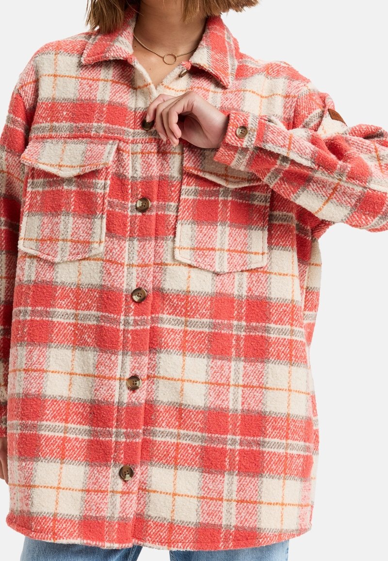 Personne portant une veste en laine à carreaux rouge, crème et orange avec boutons et deux poches poitrine, tenant un bouton près du col.