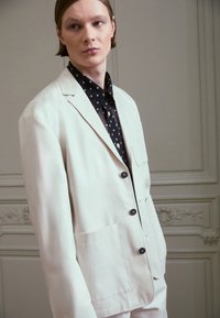 Ljusbeige blazer med två framfickor, svarta knappar, bärs över en svart blus med vita prickar. Slät textur och skräddarsydd passform.